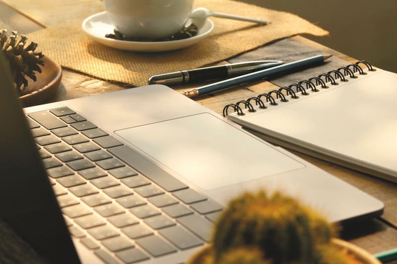 Sunlit wooden desk with a laptop, open notebook and coffee cup